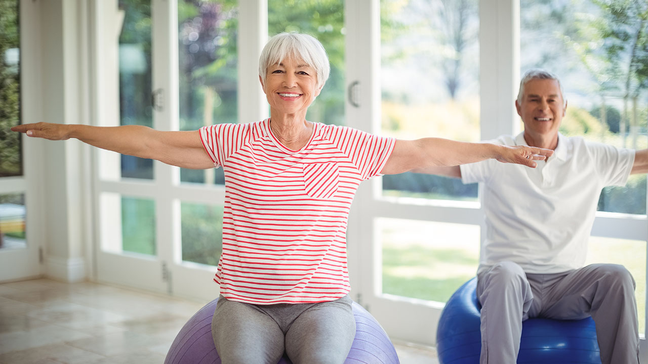 Two Seniors sit on medicine balls with their arms stretched out while smiling